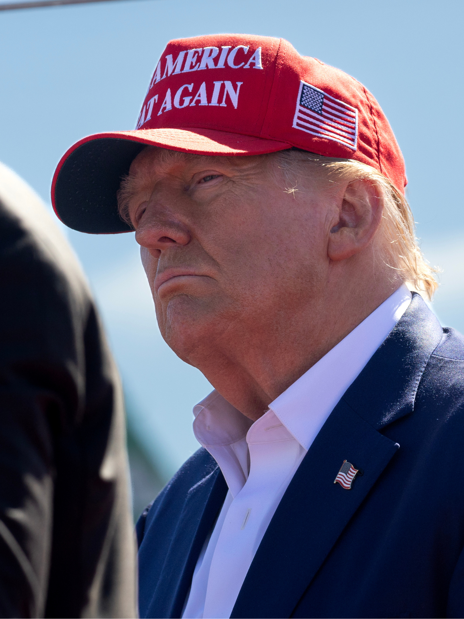 Donald Trump, in a red Make America Great Again hat, staring at someone off-camera speaking at a rally.