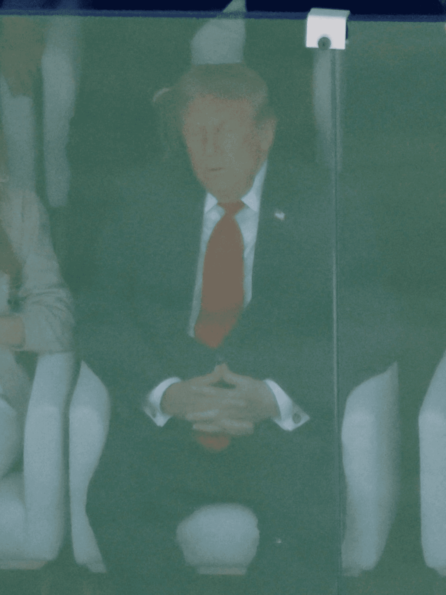 Donald Trump in the middle, with Melania Trump on the left and Gianni Infantino on the right, watching the World Cup final behind a glass screen. Trump appears to have his eyes closed.
