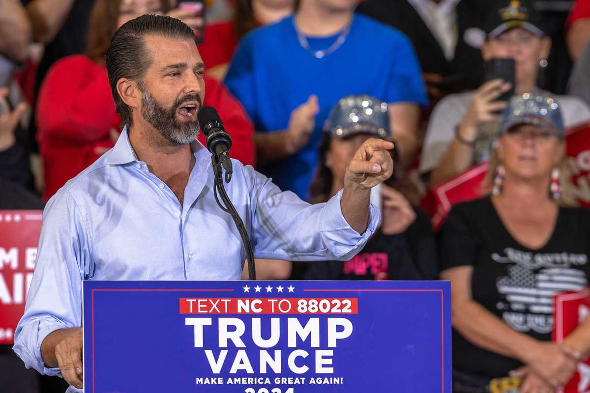 Donald Trump Jr speaking at a rally behind a lectern with blue 'Trump Vance 2024' signage on it.