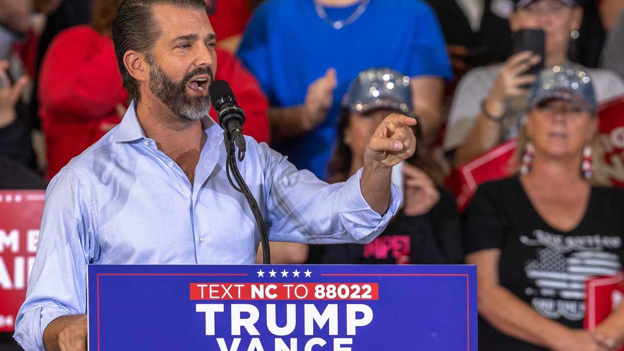 Donald Trump Jr speaking at a rally behind a lectern with blue 'Trump Vance 2024' signage on it.