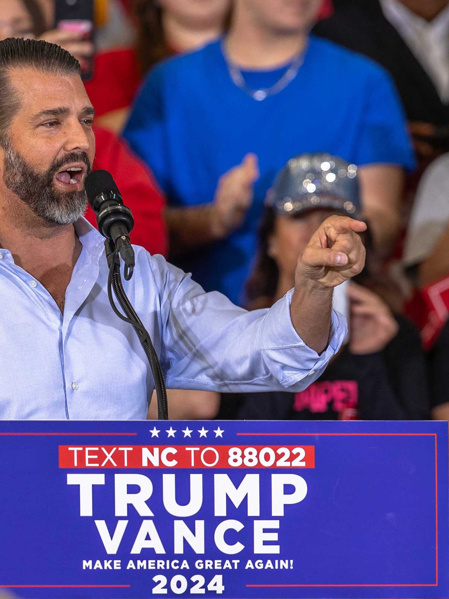 Donald Trump Jr speaking at a rally behind a lectern with blue 'Trump Vance 2024' signage on it.