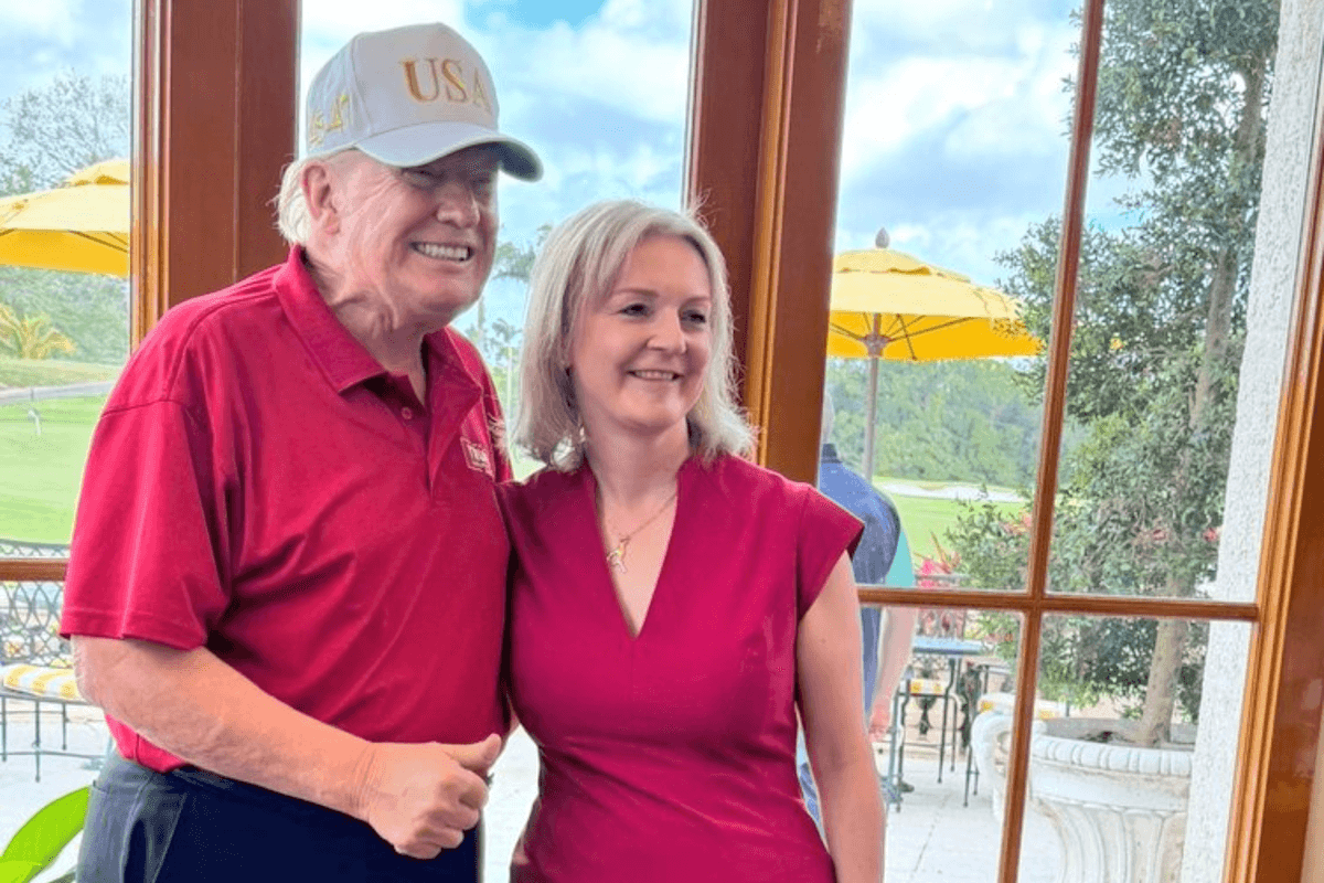 Donald Trump, left, and Liz Truss, right, smile for a photo together. Trump wears a white USA cap and a red polo shirt, while Truss wears a reddish pink dress.