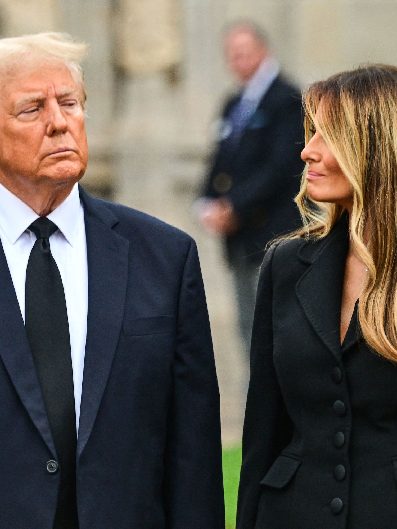 Donald Trump, left, looks ahead with a frowning facial expression, while Melania, right, with wavy blonde hair, turns her head right to look at him with a faint smile.