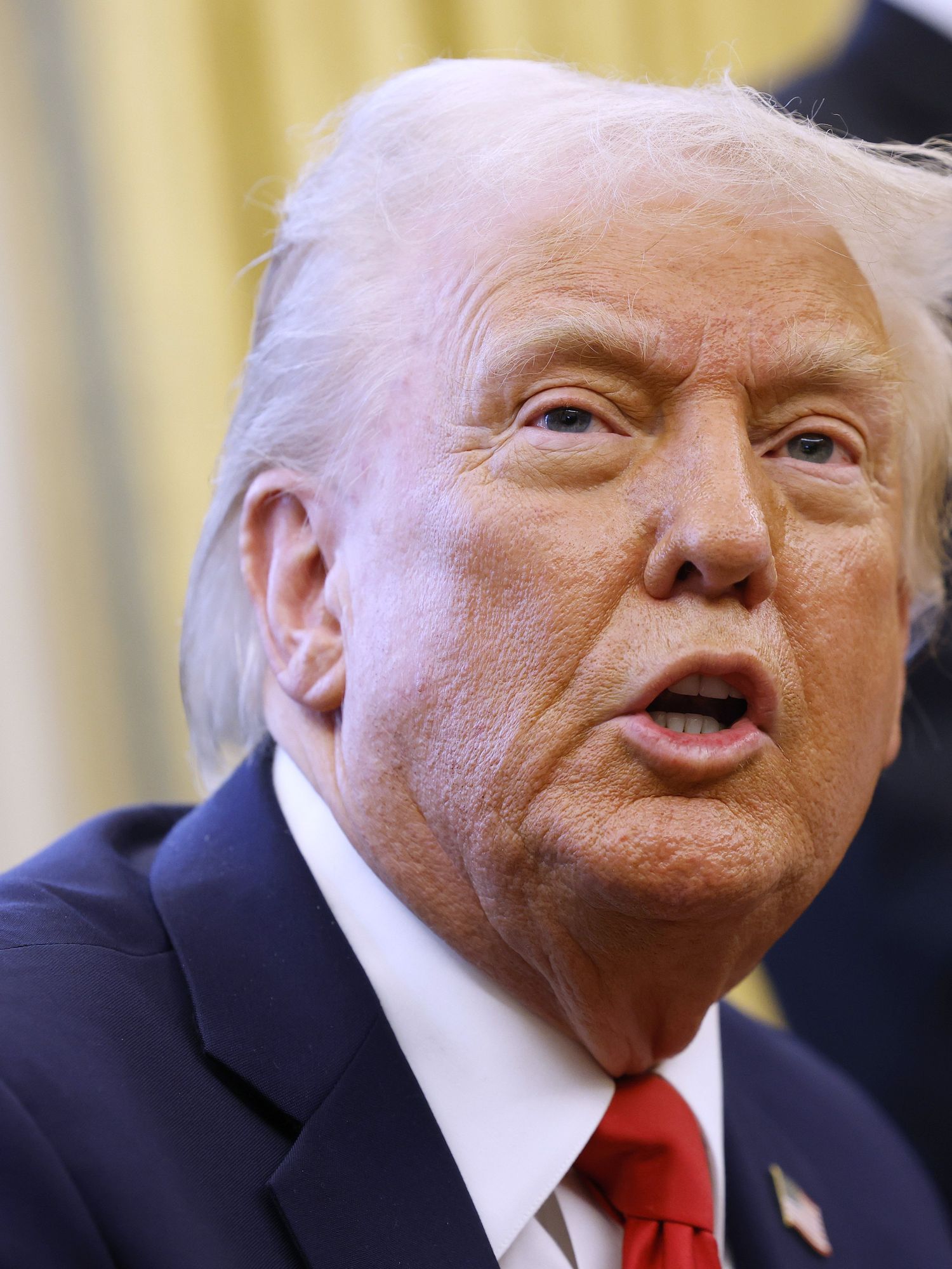 Donald Trump looks at someone off-camera as he sits in the Oval Office, wearing a dark blue suit and red tie.