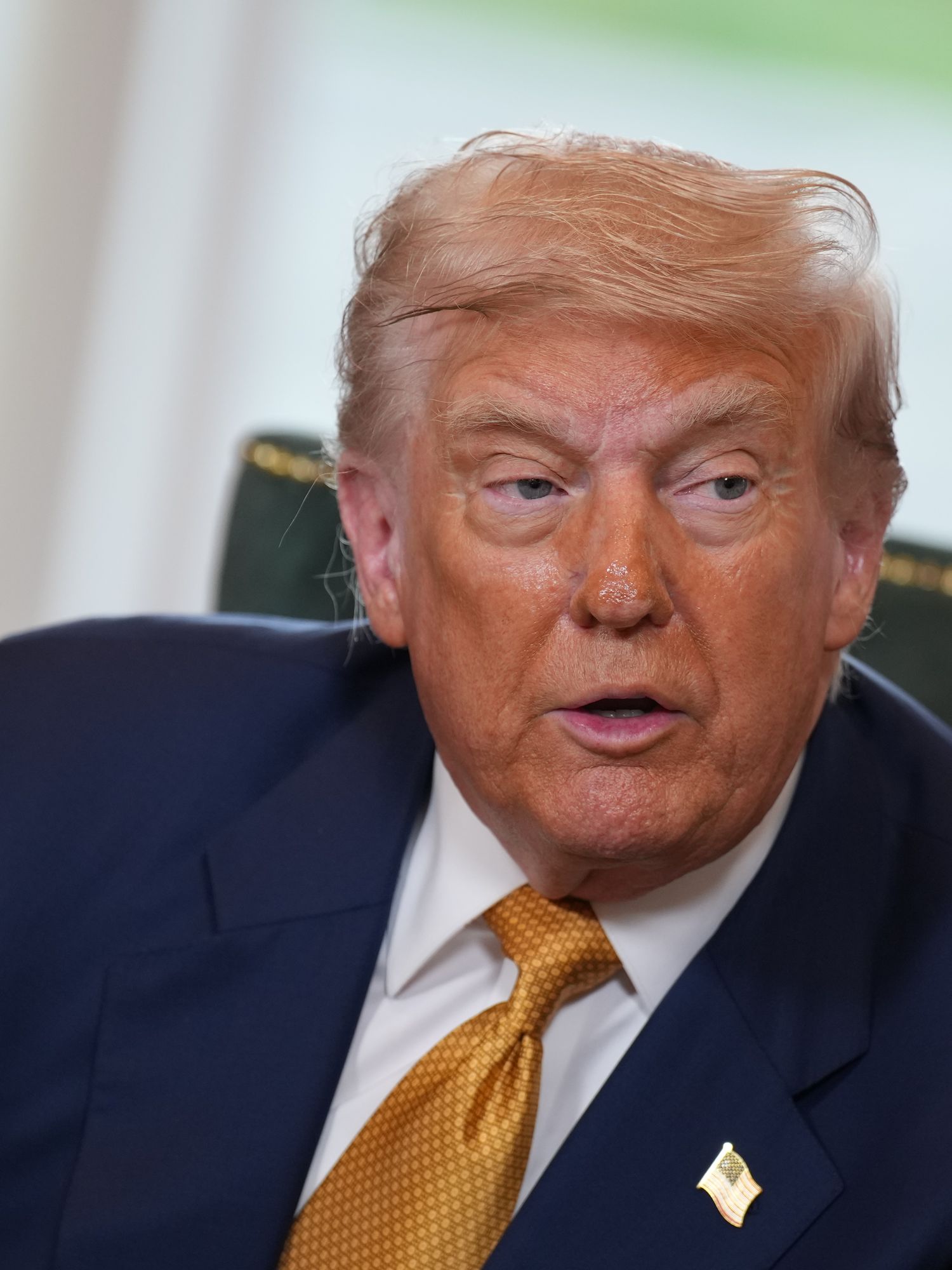 Donald Trump looks to his left as he sits on a green chair. He's wearing a blue suit and yellow tie.