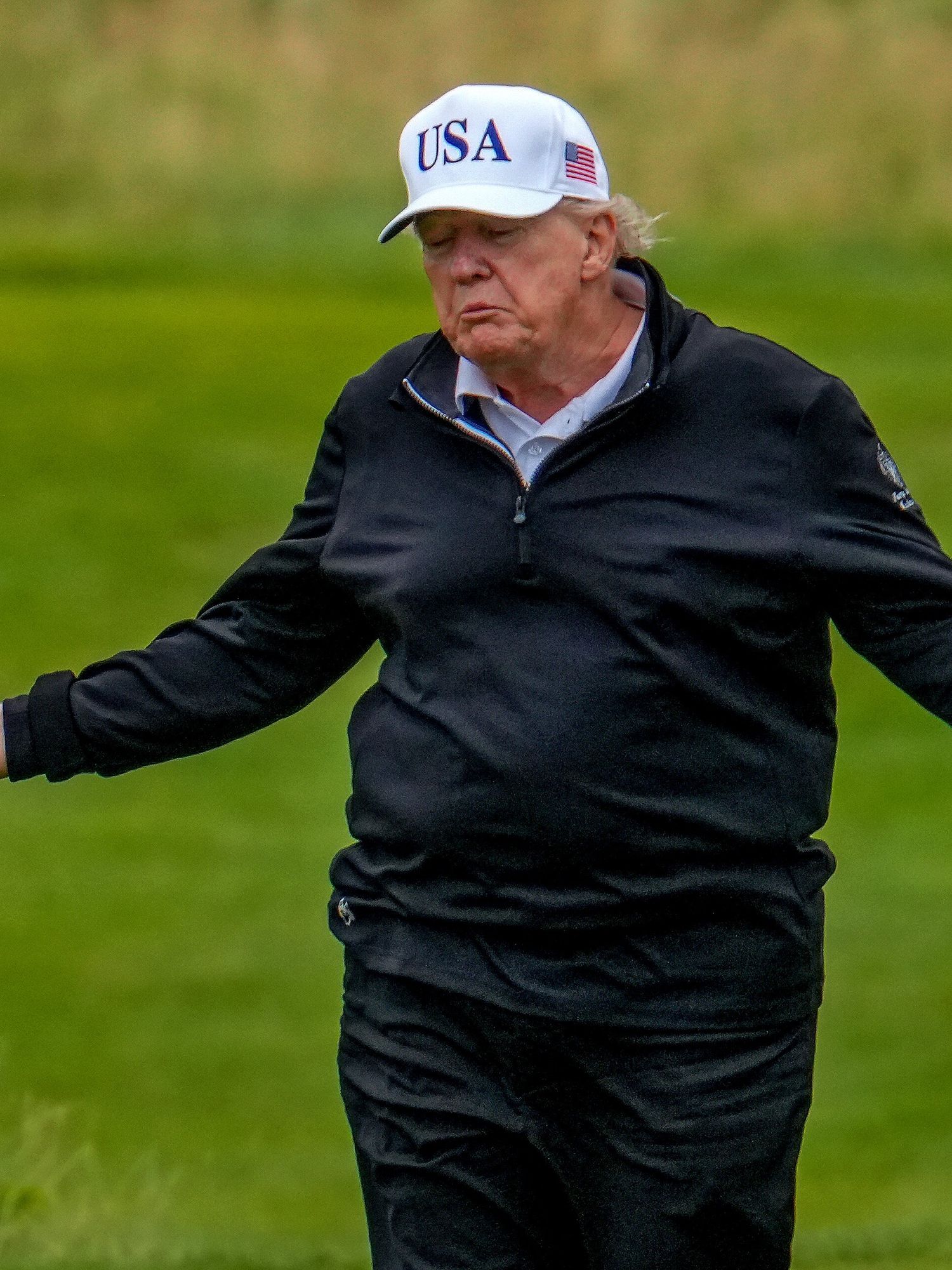 Donald Trump on a golf course in a black jacket and white USA cap, shrugging his shoulders with his arms out wide.