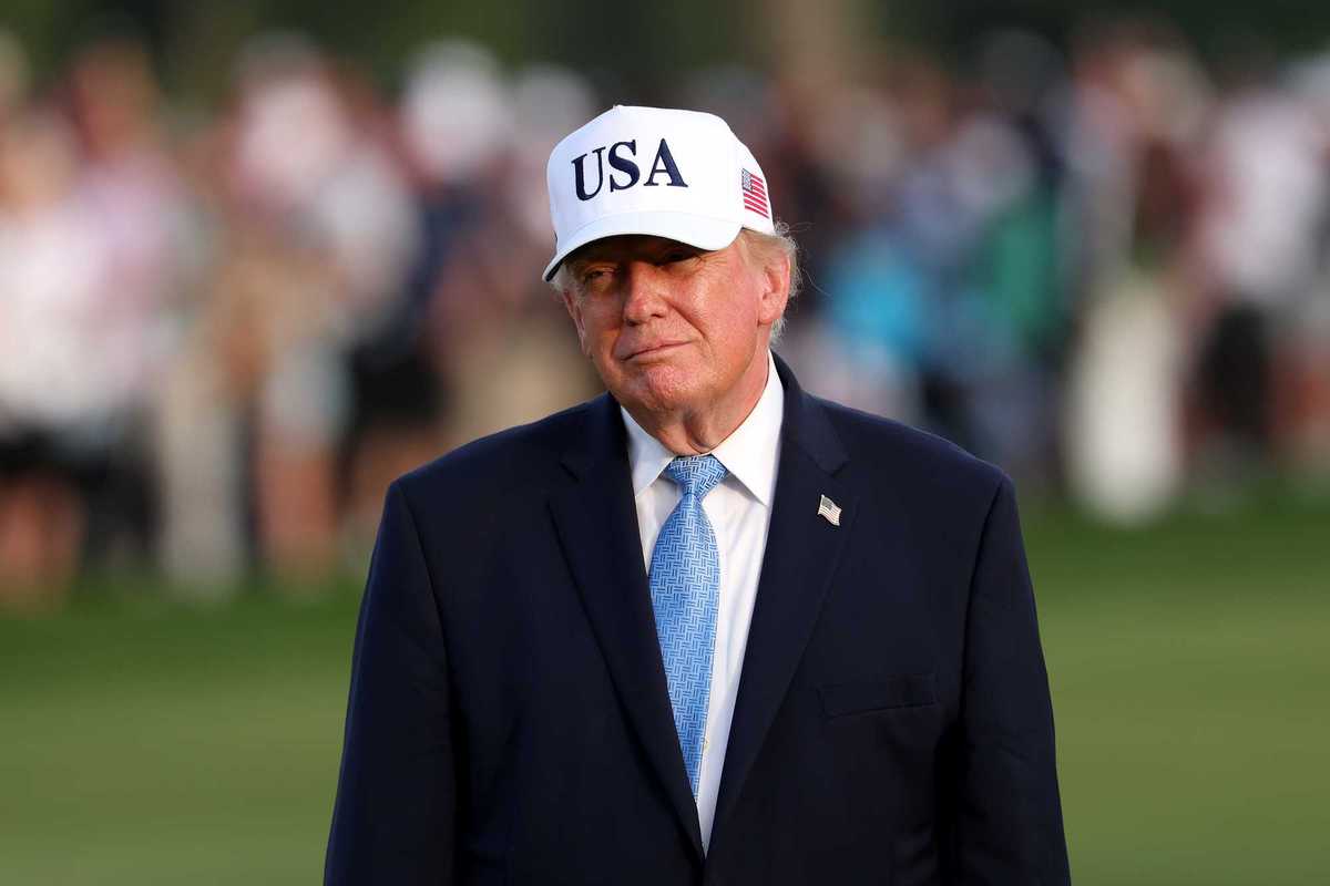 Donald Trump on a golf course, wearing a black suit and white USA cap.