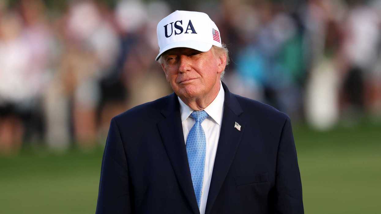 Donald Trump on a golf course, wearing a black suit and white USA cap.