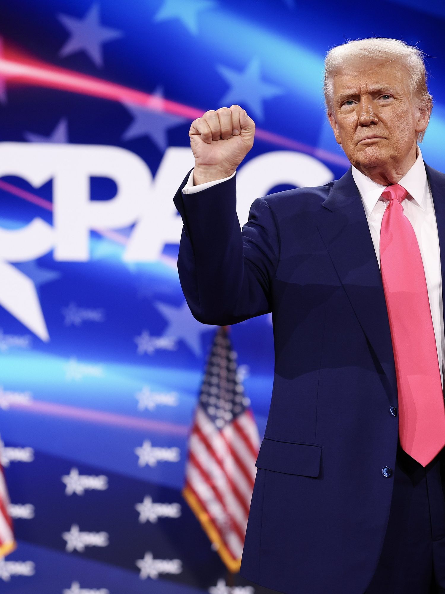 Donald Trump on stage at CPAC, wearing a dark blue suit, frowning, and raising his right fist up in a 'solidarity' gesture.