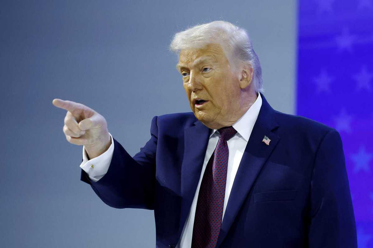 Donald Trump points with his right index finger at something off-camera. His face is visibly covered in makeup.
