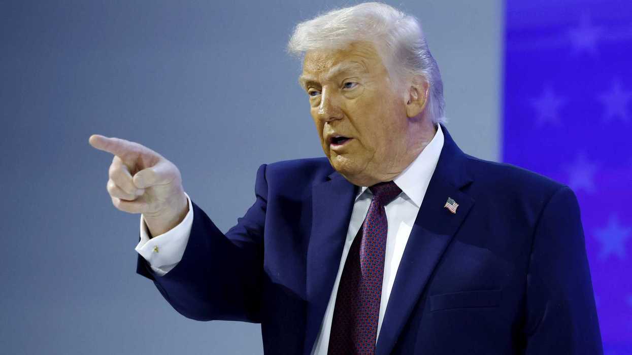 Donald Trump points with his right index finger at something off-camera. His face is visibly covered in makeup.