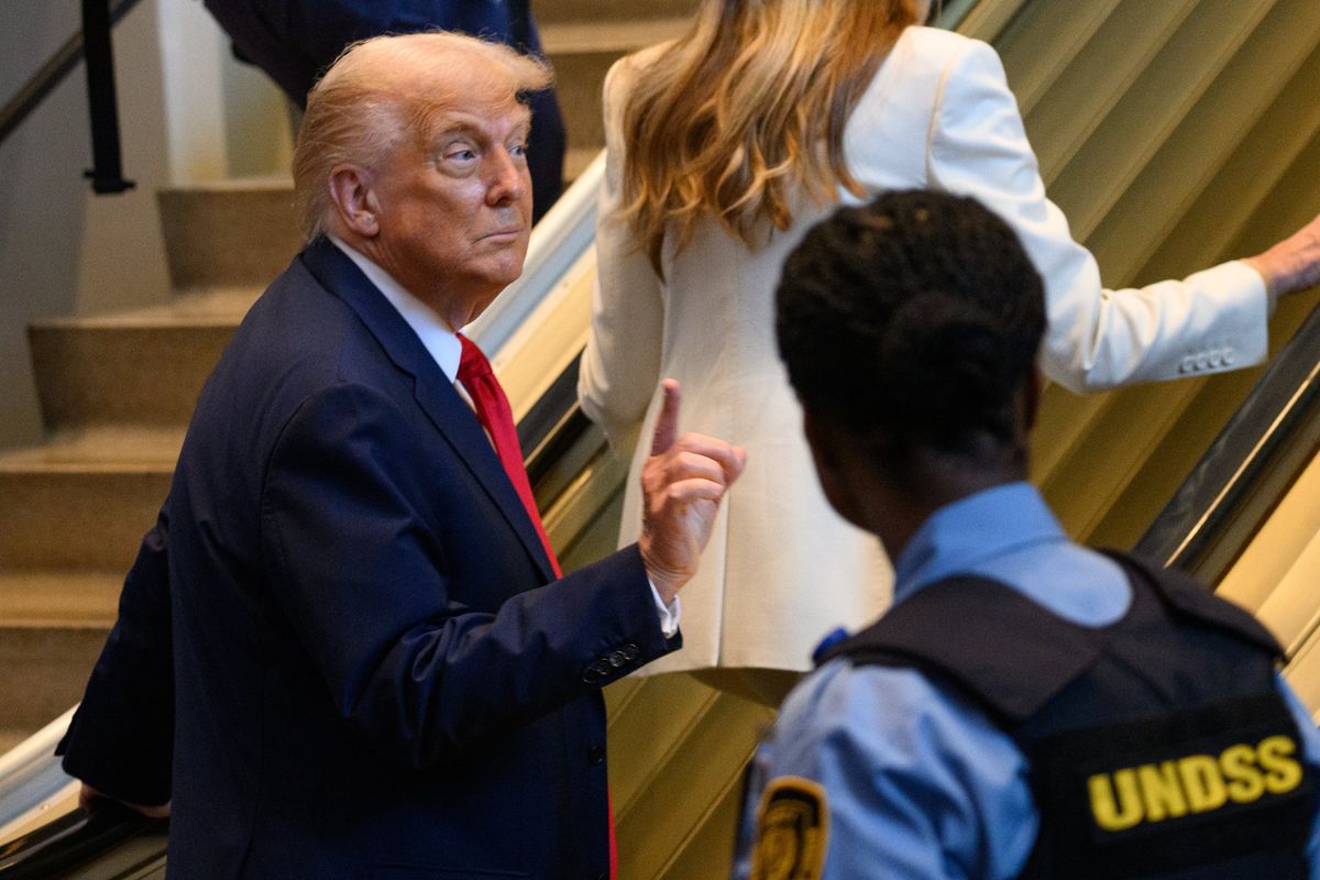 Donald Trump raises his right index finger, looking to someone off-camera on his right, as he walks up an escalator.