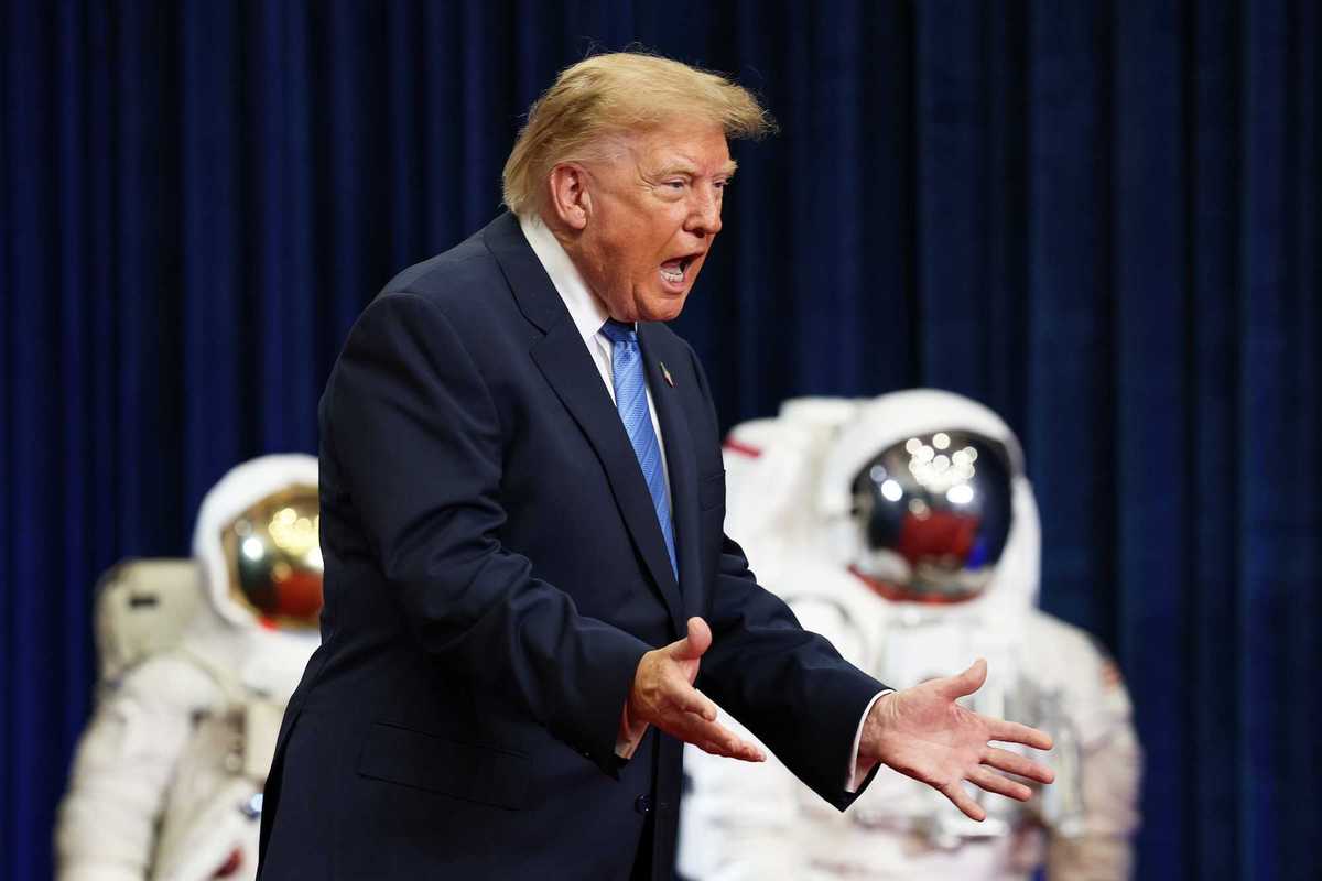 Donald Trump shouts with his arms out in front of him. In the background are two spacesuits.