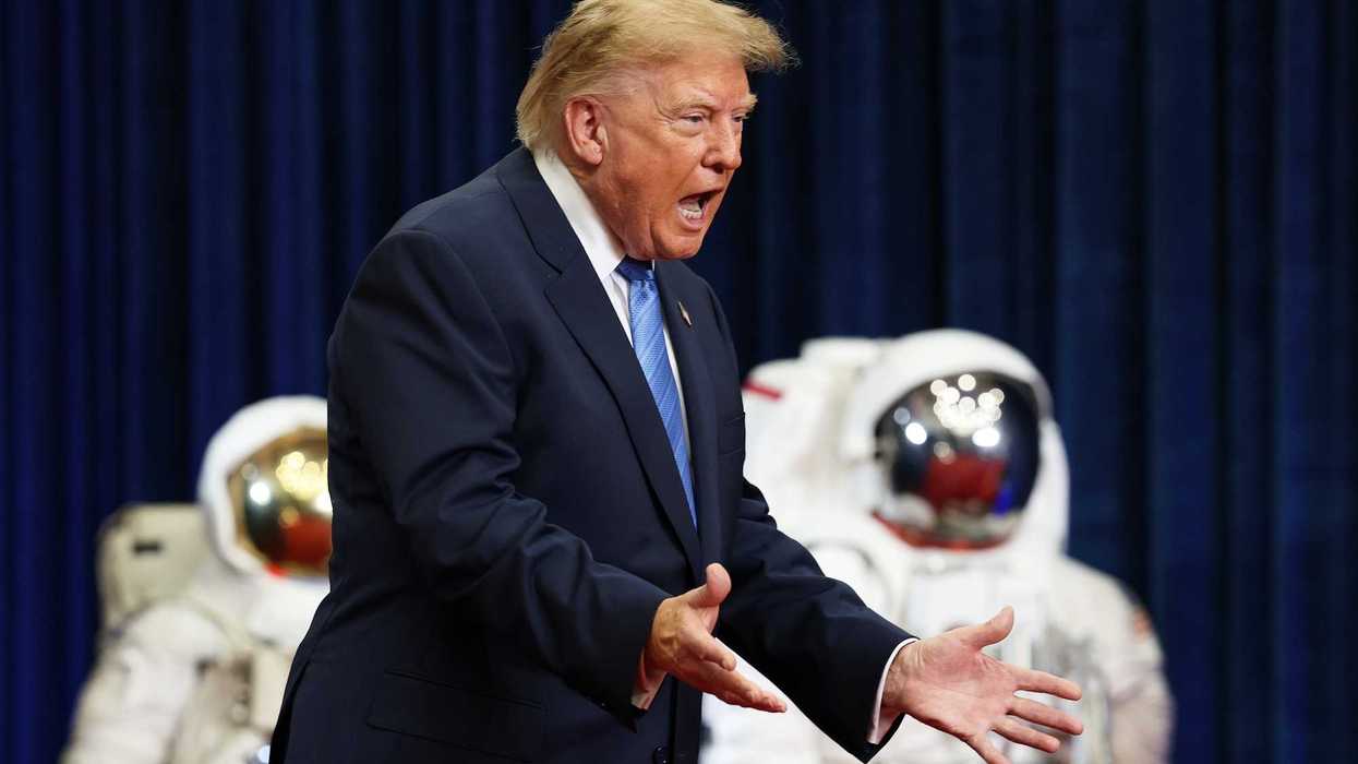 Donald Trump shouts with his arms out in front of him. In the background are two spacesuits.