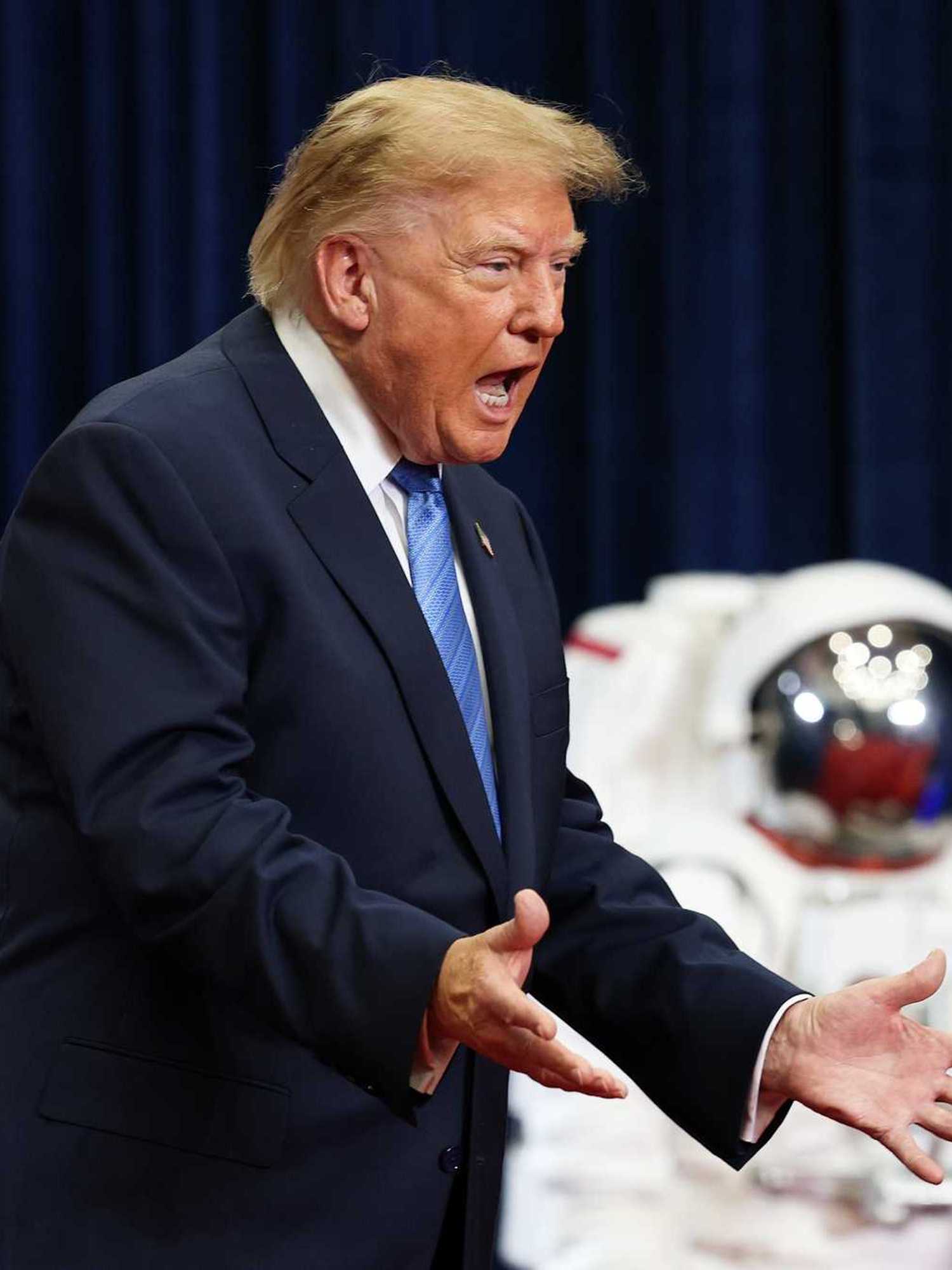 Donald Trump shouts with his arms out in front of him. In the background are two spacesuits.
