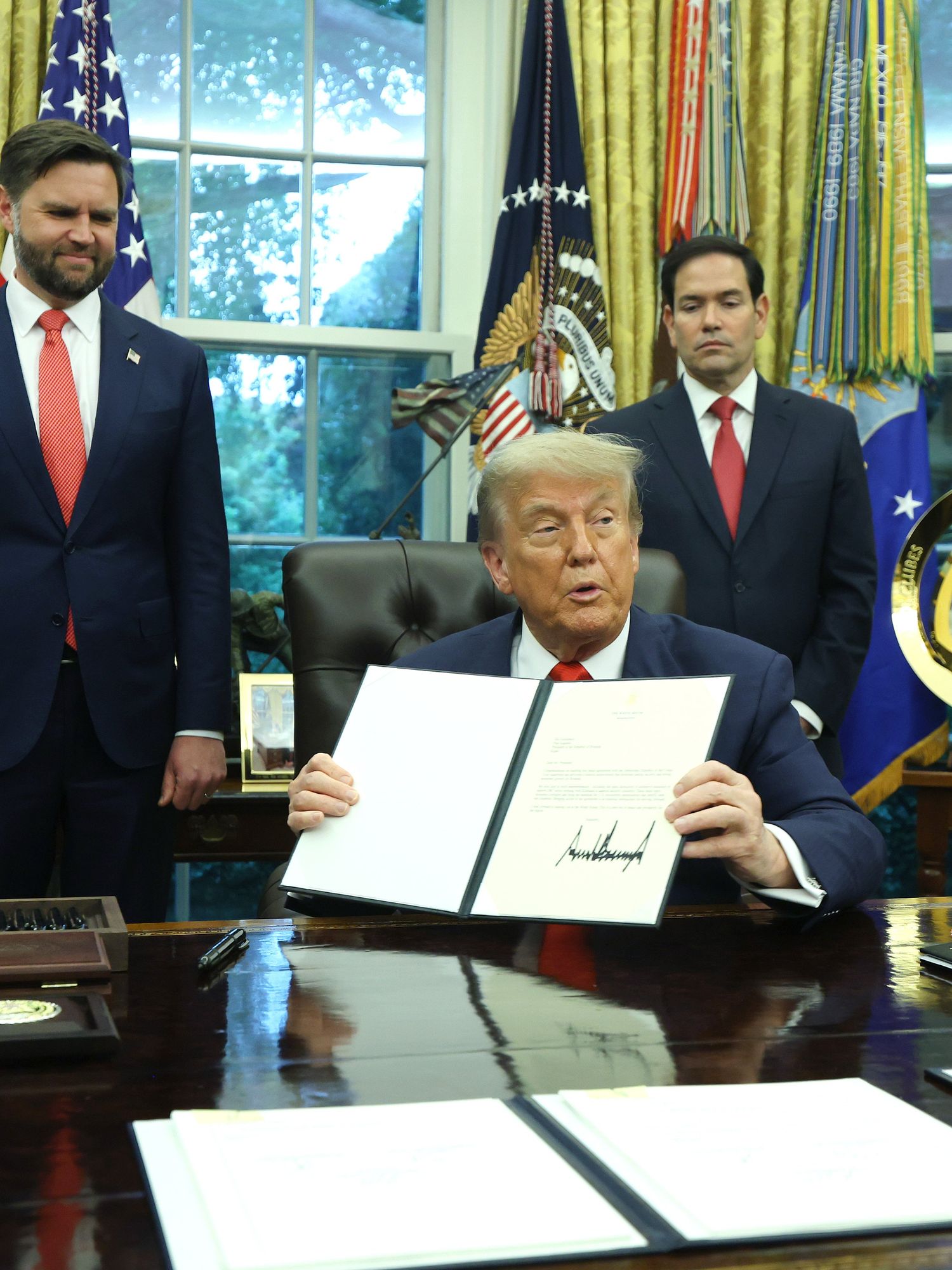 Donald Trump shows a signed document to the press behind his Oval Office desk, with four people standing behind him, looking at him.