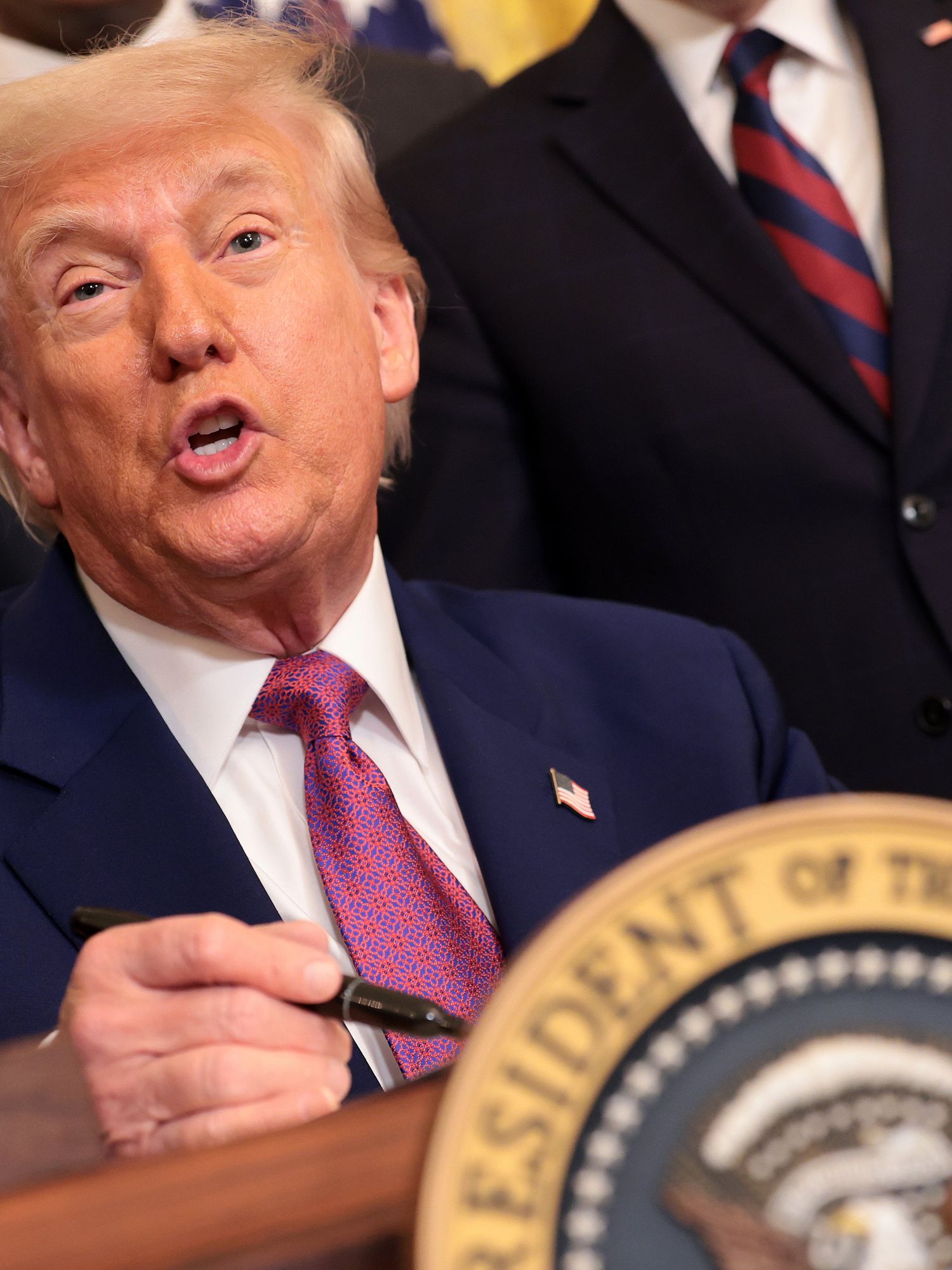 Donald Trump signing an executive order behind a wooden desk with the presidential emblem on it, talking to reporters.