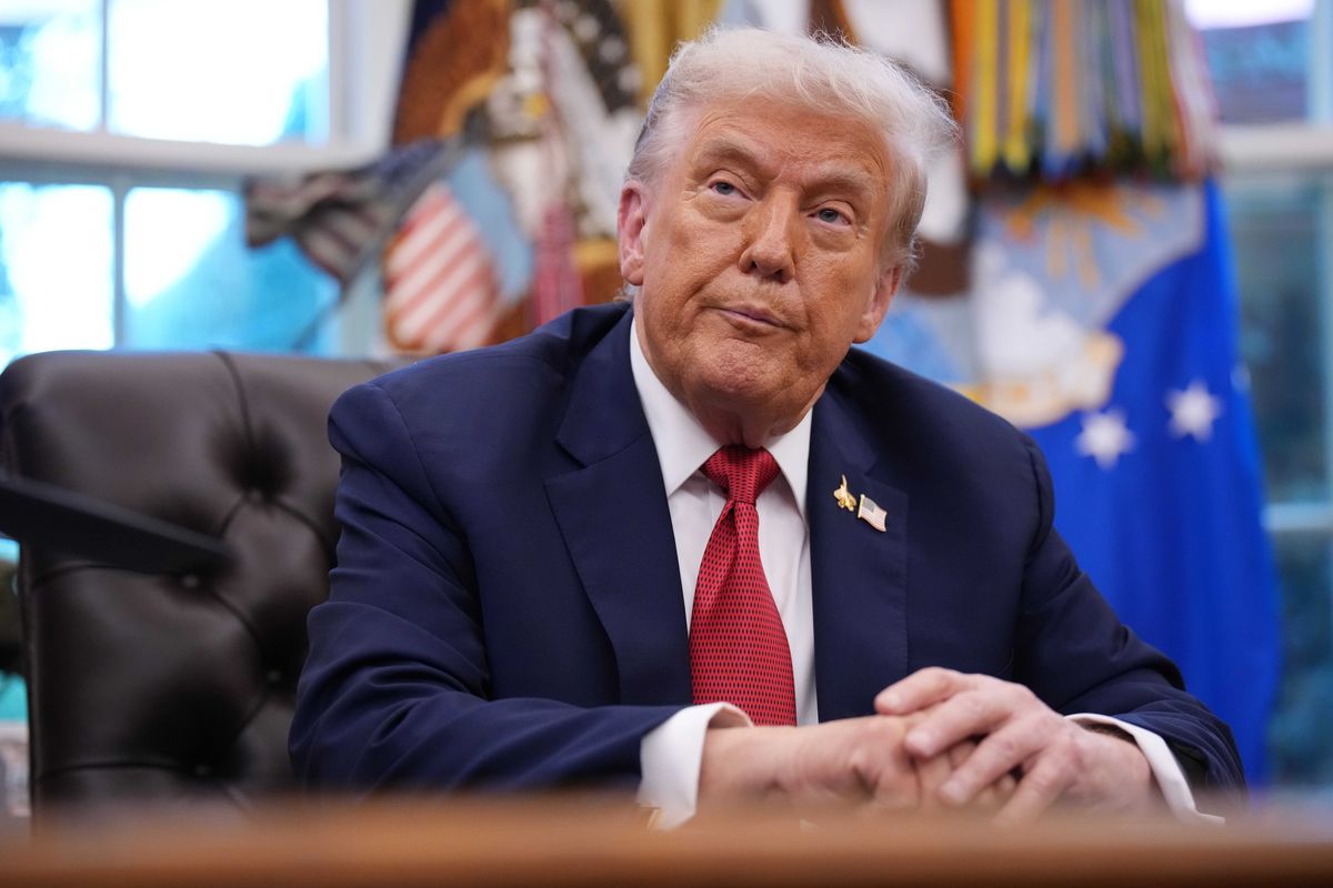 Donald Trump sits at the Oval Office desk, turning and tilting his head slightly to look at something on his right.