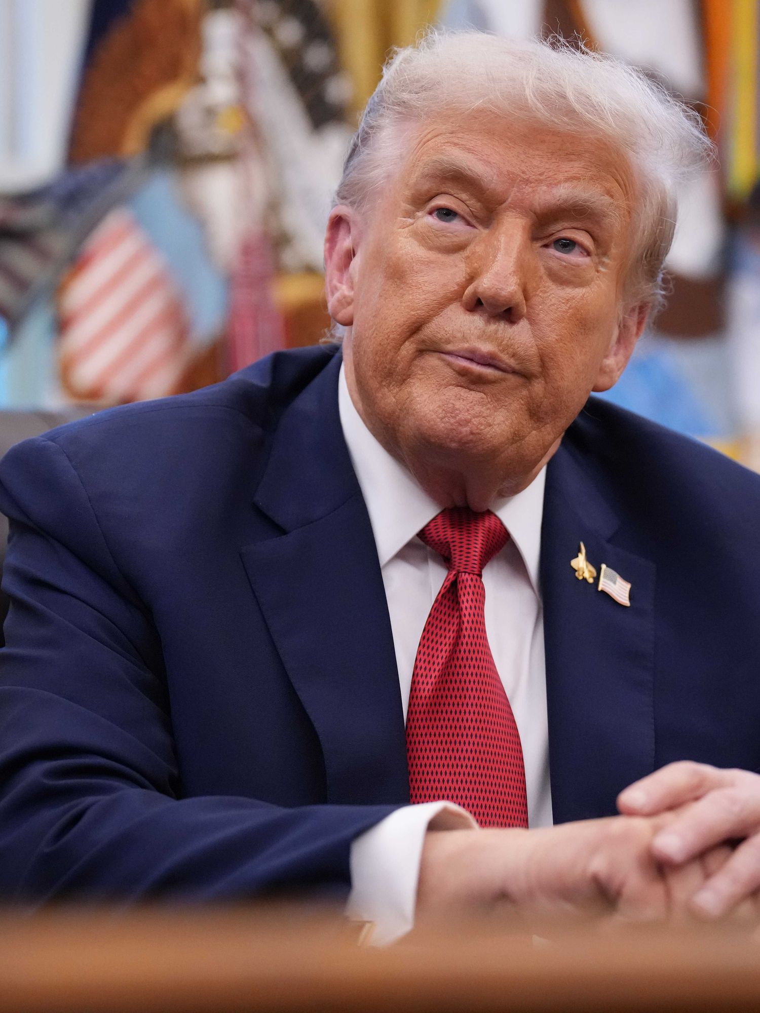 Donald Trump sits at the Oval Office desk, turning and tilting his head slightly to look at something on his right.