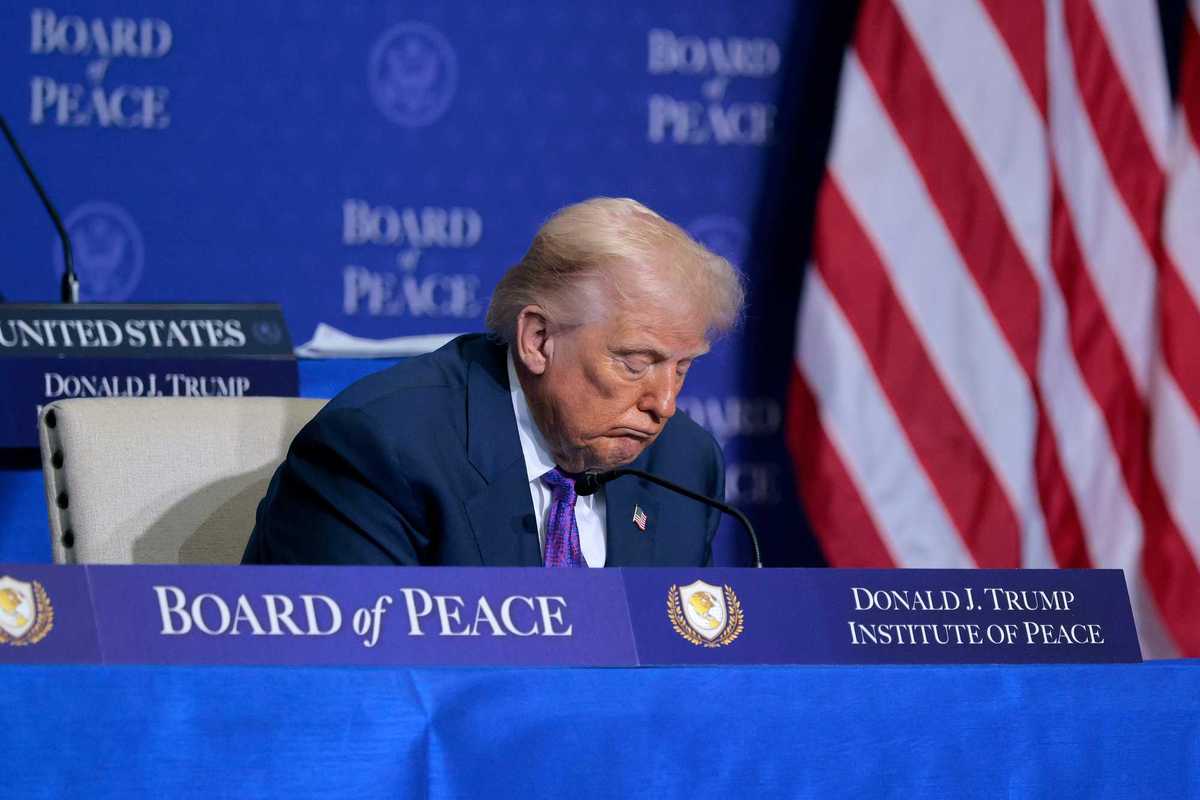 Donald Trump sitting behind a blue desk with 'Board of Peace' and 'Donald J. Trump Institute of Peace' signage on it. Trump has his head lowered, and appears to be asleep.
