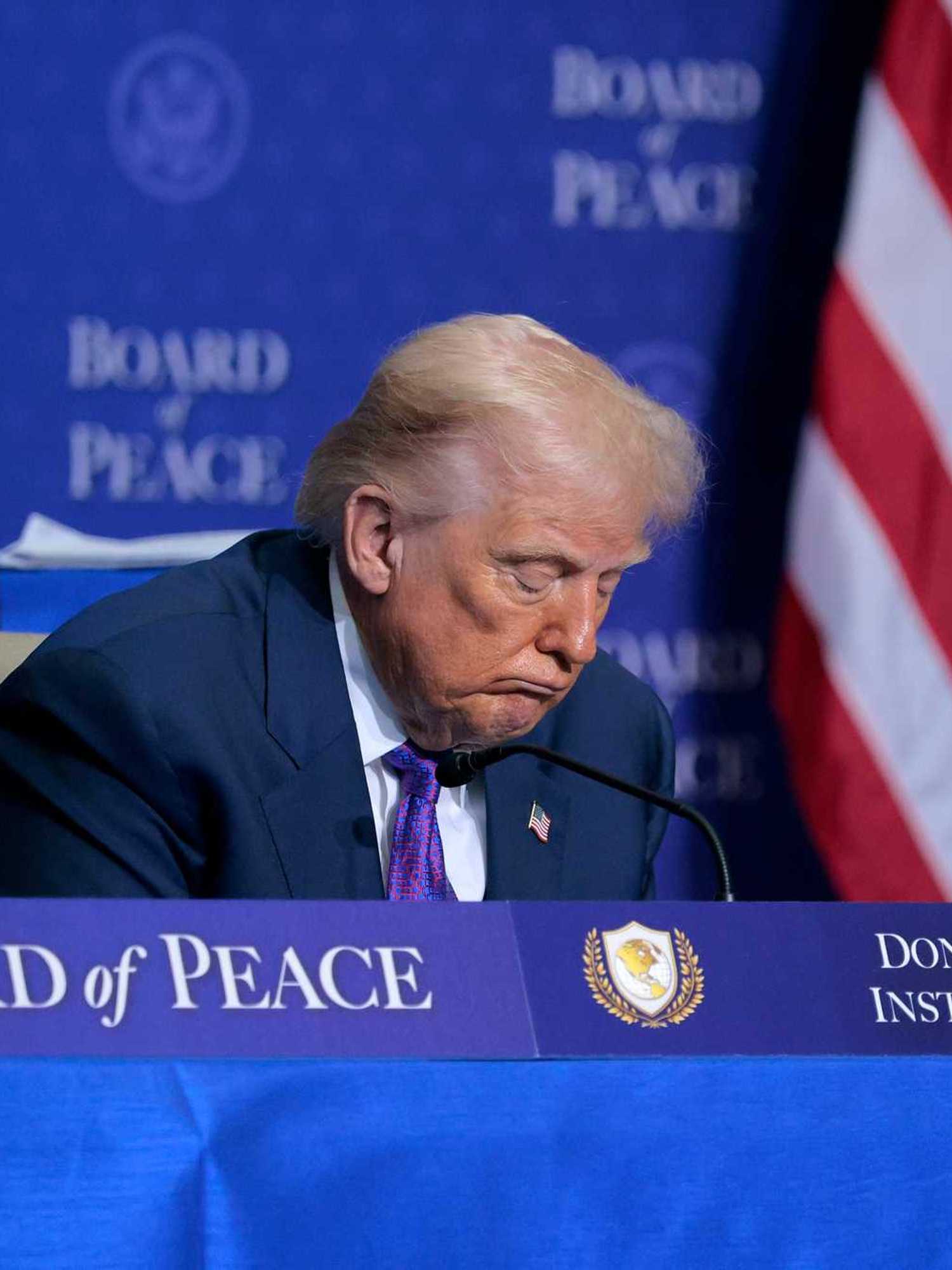 Donald Trump sitting behind a blue desk with 'Board of Peace' and 'Donald J. Trump Institute of Peace' signage on it. Trump has his head lowered, and appears to be asleep.