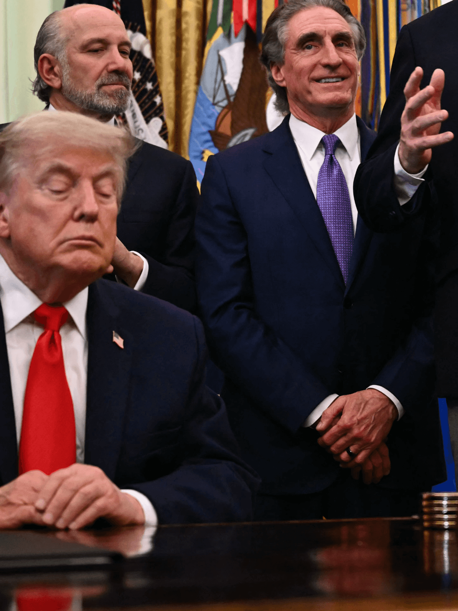 Donald Trump sitting behind the Resolute Desk in the Oval Office, appearing to have his eyes closed.