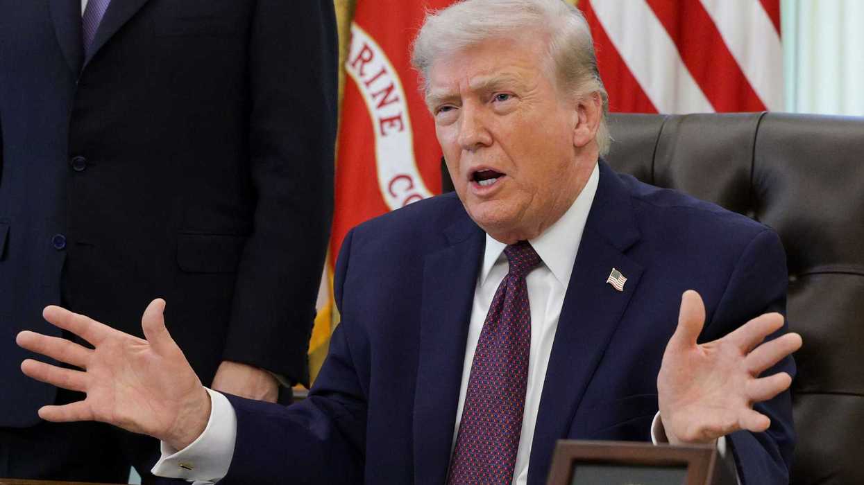 Donald Trump sitting behind the Resolute Desk in the Oval Office with his arms spread wide in front of him.
