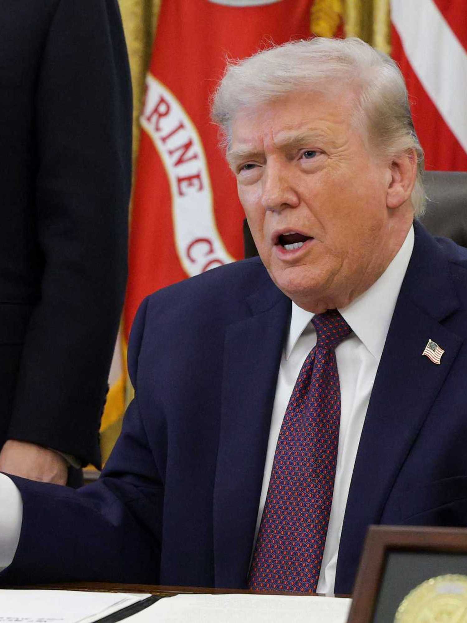 Donald Trump sitting behind the Resolute Desk in the Oval Office with his arms spread wide in front of him.