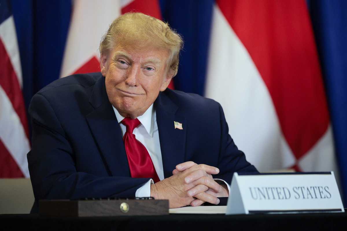 Donald Trump smiles as he sits at a table with a sign saying 'United States' on it.
