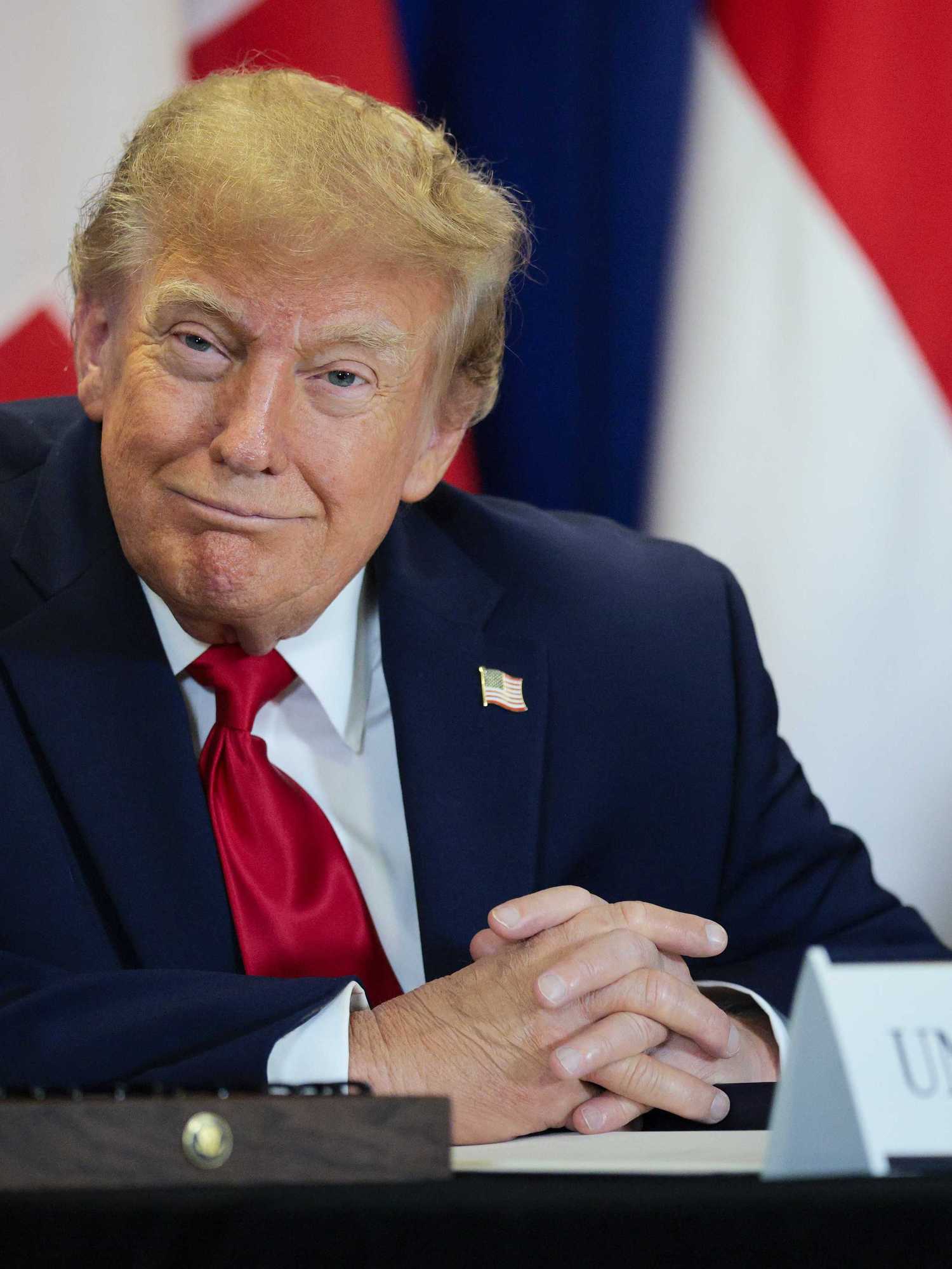 Donald Trump smiles as he sits at a table with a sign saying 'United States' on it.