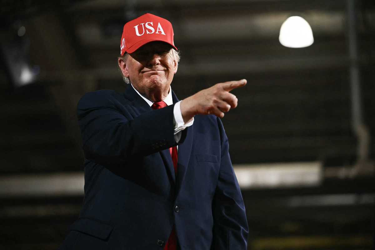 Donald Trump smiling and pointing with his right index finger. He's wearing a red USA cap.