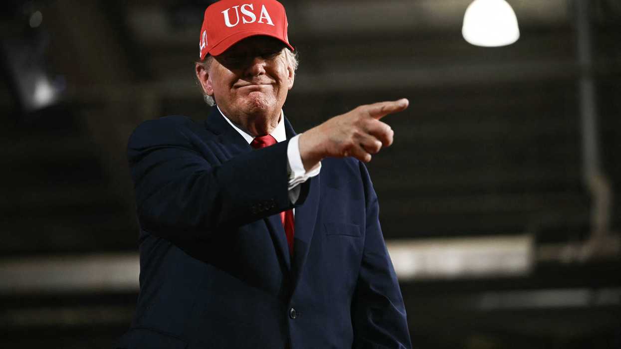 Donald Trump smiling and pointing with his right index finger. He's wearing a red USA cap.