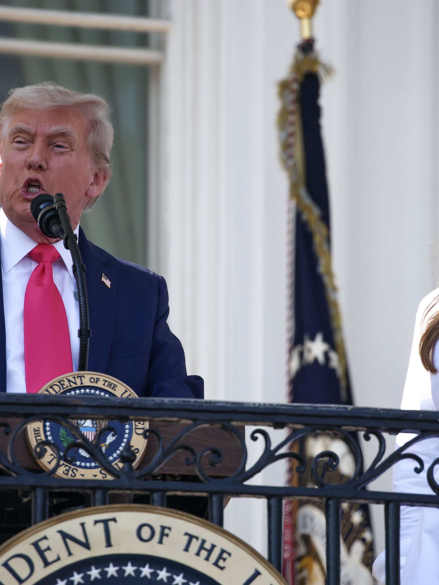 Donald Trump speaks behind a lectern on a White House balcony, with Melania standing to his left.
