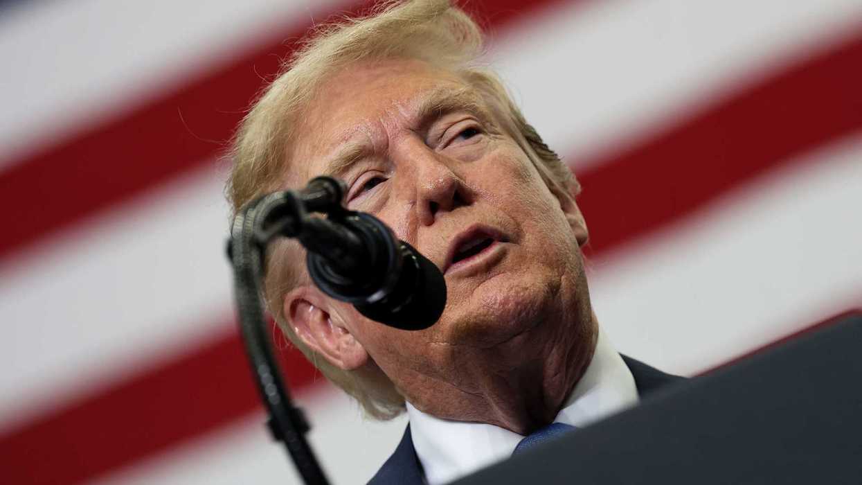 Donald Trump speaks into a microphone. A giant American flag is out of focus behind him.