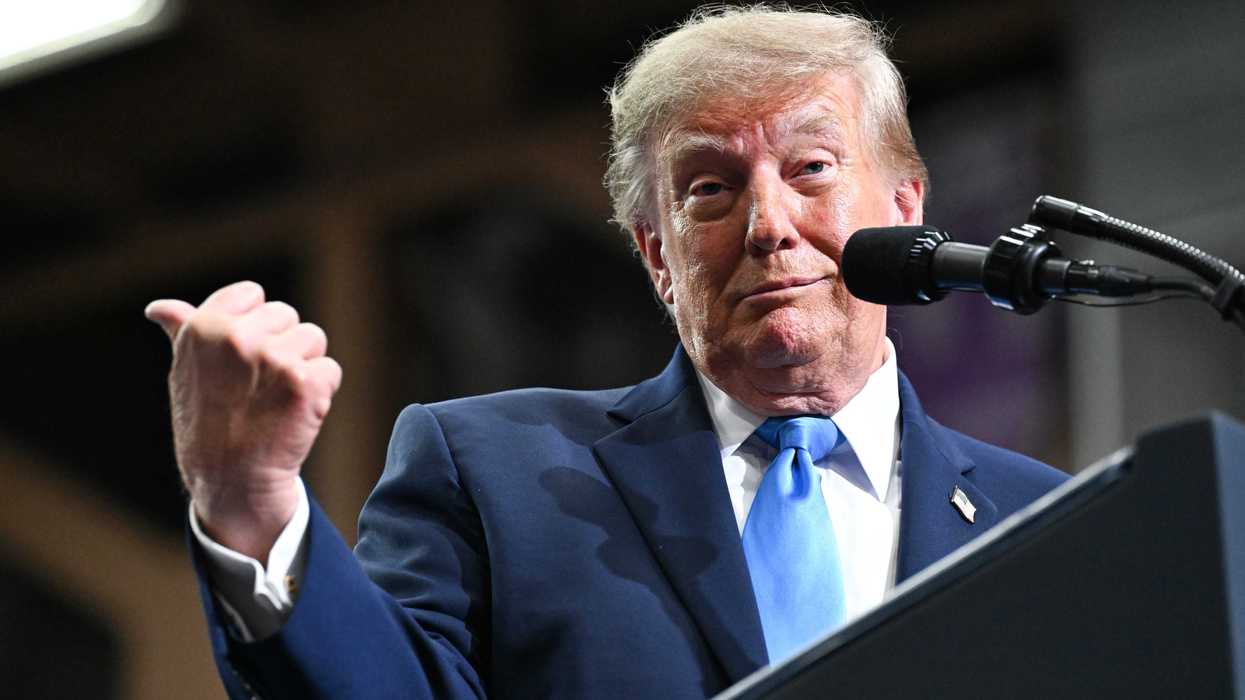 Donald Trump standing behind a lectern and pointing behind him with his right thumb, smirking.