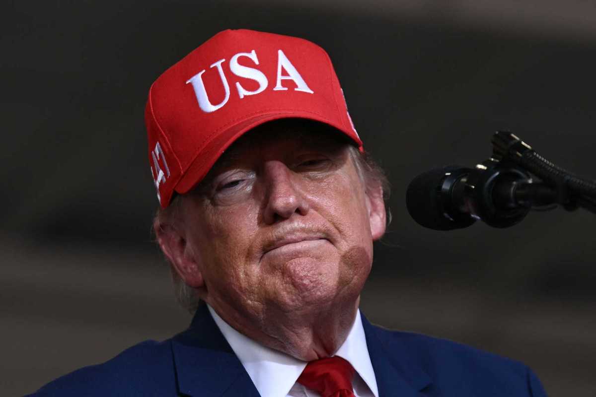 Donald Trump standing behind a microphone wearing a red USA cap and blue suit.