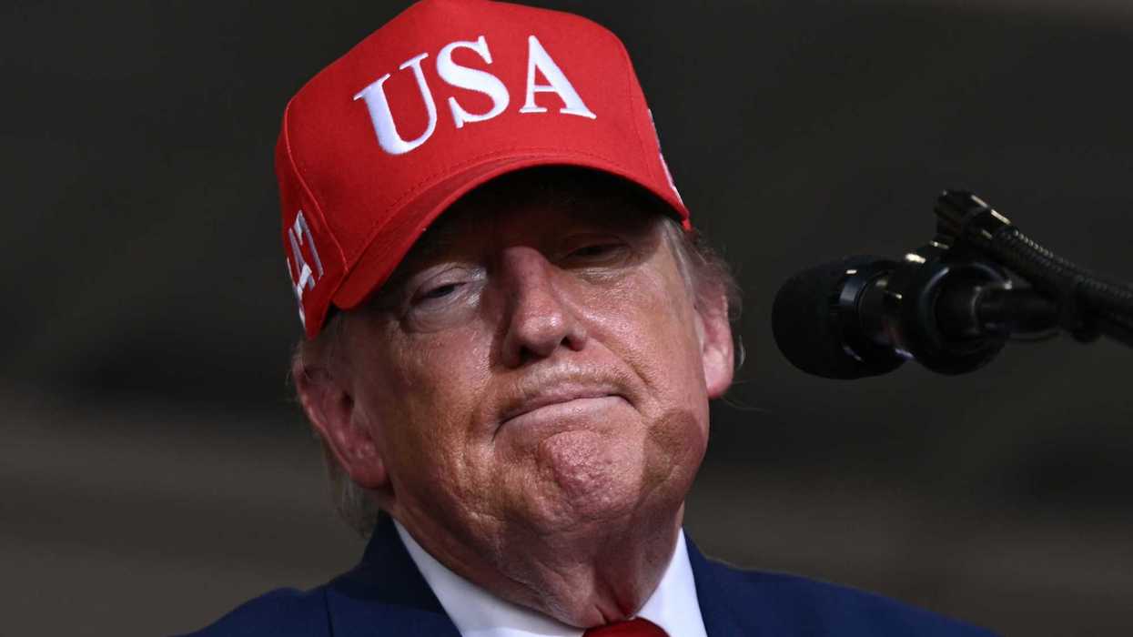 Donald Trump standing behind a microphone wearing a red USA cap and blue suit.