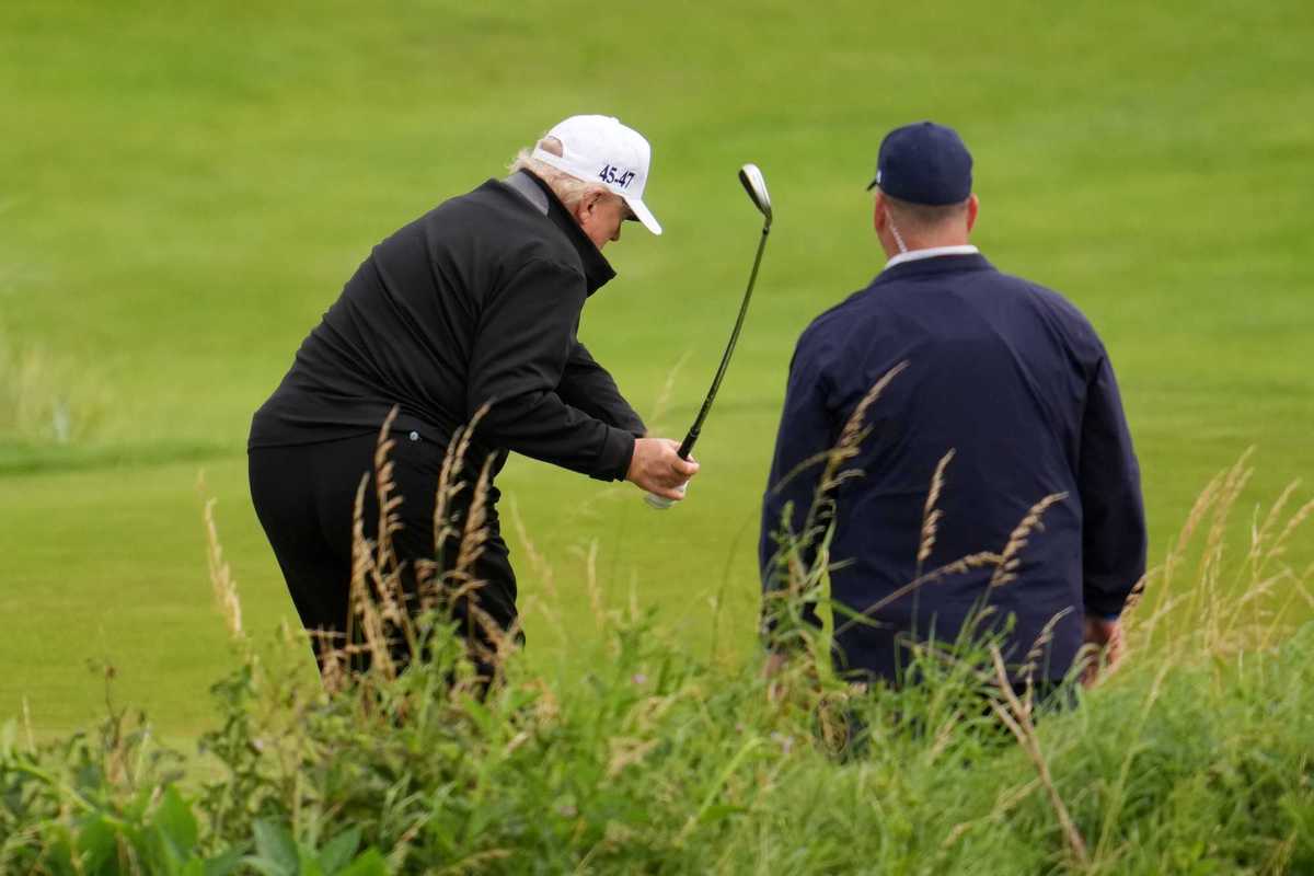 Donald Trump swings a golf club on a golf course, with a white man with his back to the camera watching on his right.