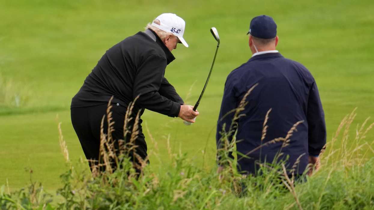 Donald Trump swings a golf club on a golf course, with a white man with his back to the camera watching on his right.