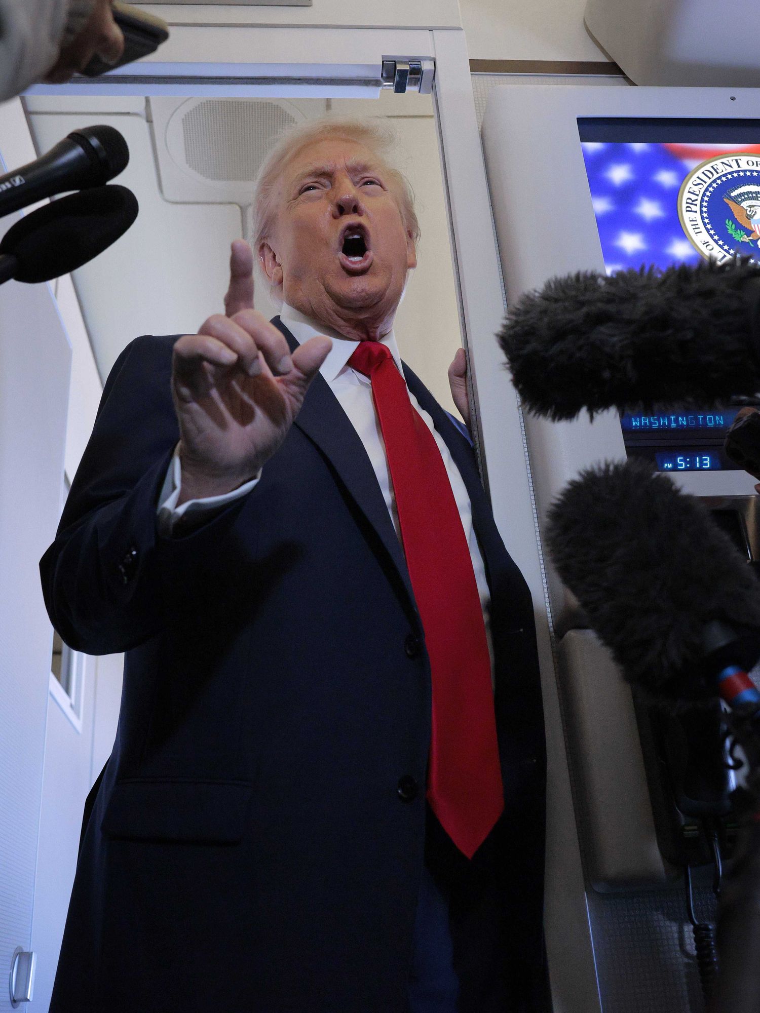 Donald Trump talks to reporters inside Air Force One, pointing upwards with his right index finger while talking in the doorway.