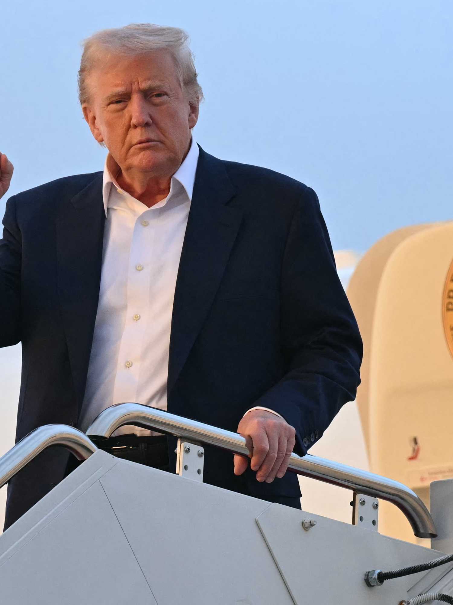 Donald Trump walks down the stairs from Air Force One, wearing a black suit and white shirt and raises his right fist defiantly.