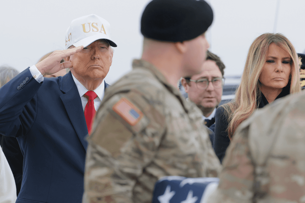 Donald Trump, wearing a blue suit and white USA cap, salutes a casket draped in the US flag and carried by soldiers during a dignified transfer.