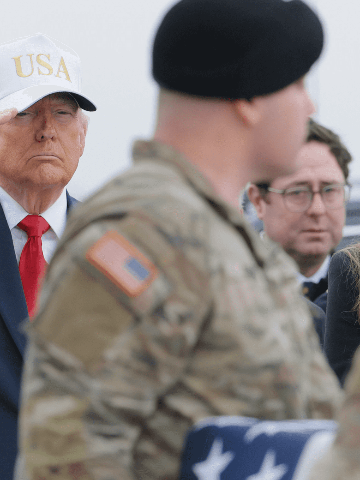 Donald Trump, wearing a blue suit and white USA cap, salutes a casket draped in the US flag and carried by soldiers during a dignified transfer.