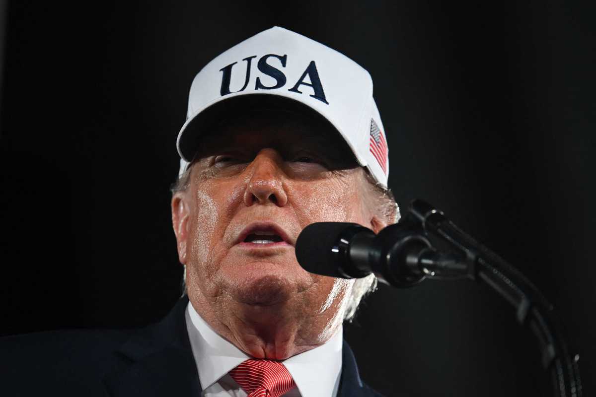 Donald Trump, wearing a white USA hat, speaks into a microphone.