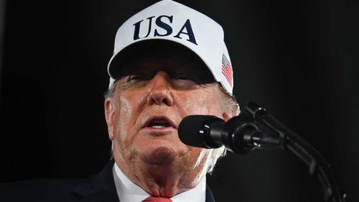 Donald Trump, wearing a white USA hat, speaks into a microphone.