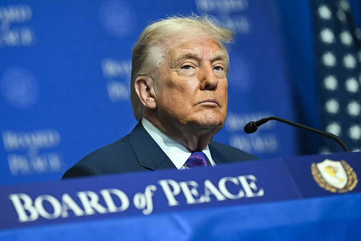 Donald Trump with a neutral expression on his face, sitting at a table with a blue 'Board of Peace' sign on it.