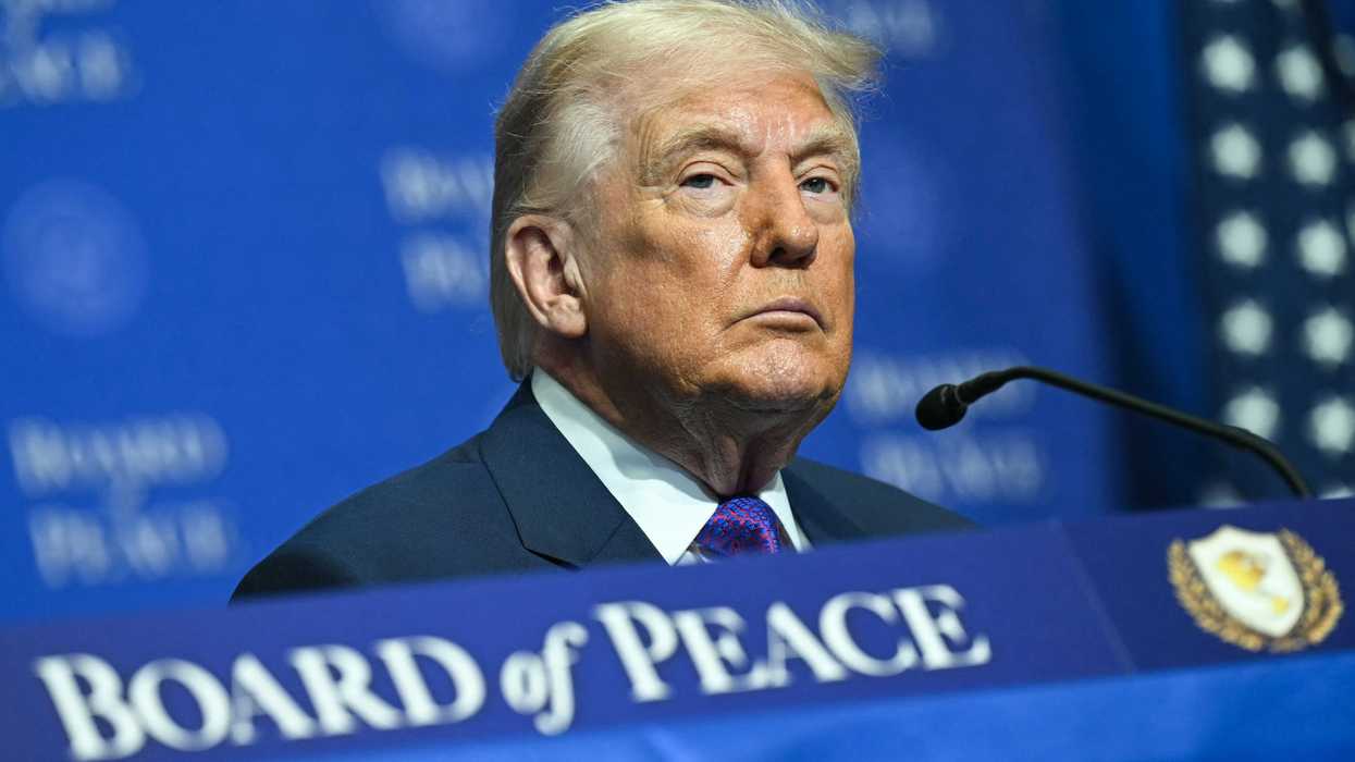 Donald Trump with a neutral expression on his face, sitting at a table with a blue 'Board of Peace' sign on it.