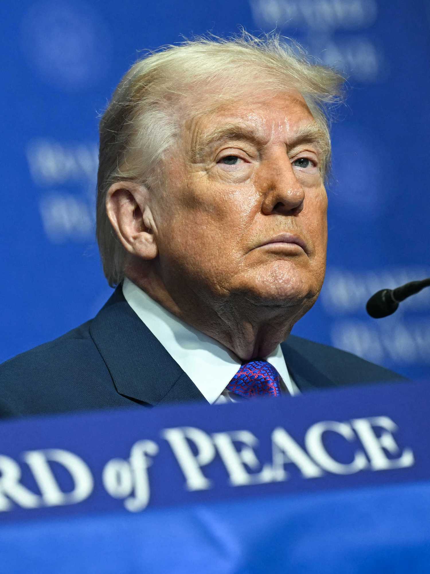 Donald Trump with a neutral expression on his face, sitting at a table with a blue 'Board of Peace' sign on it.