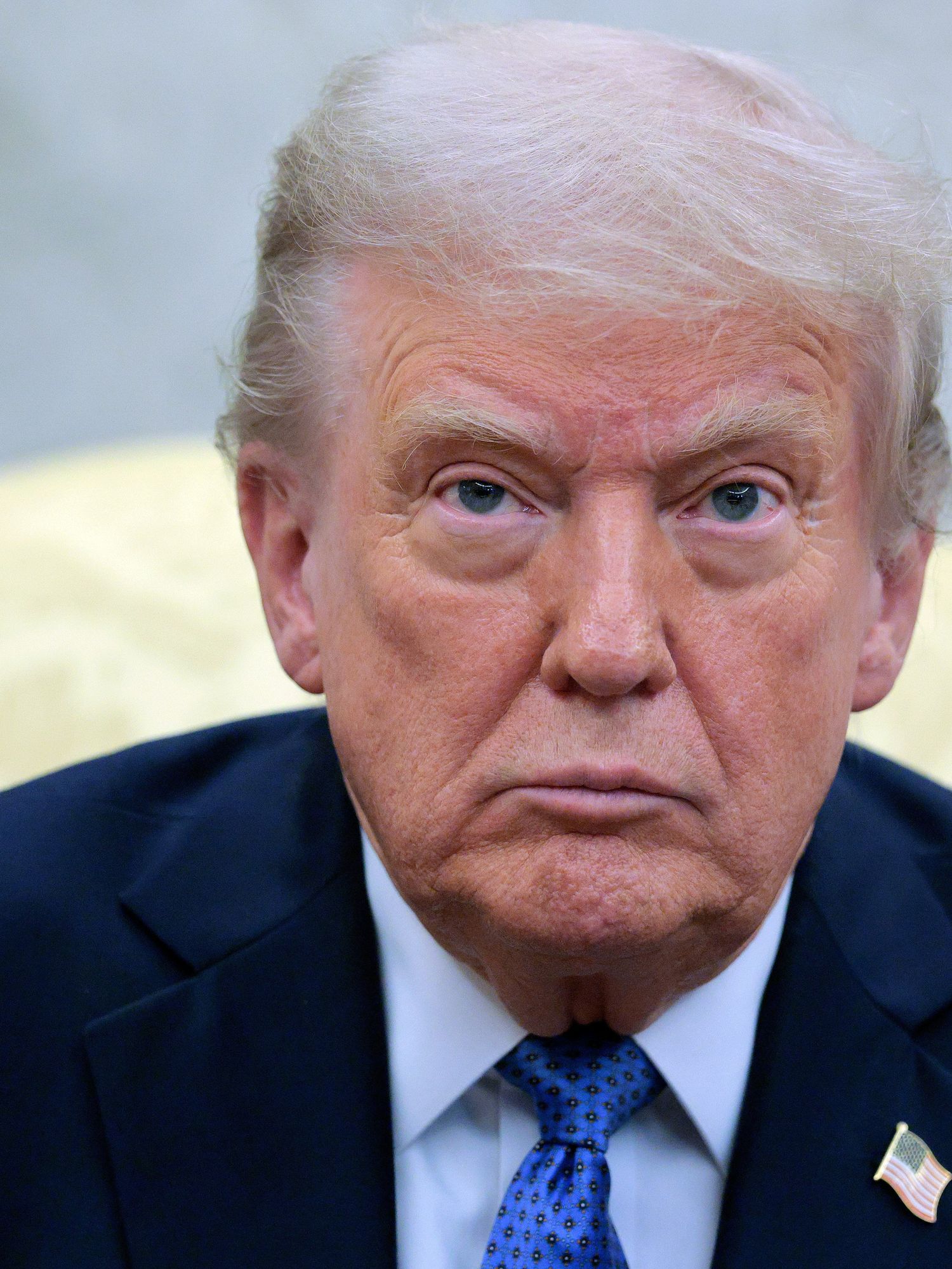 Donald Trump, with a neutral expression on his face, sitting in a light yellow chair in the Oval Office.