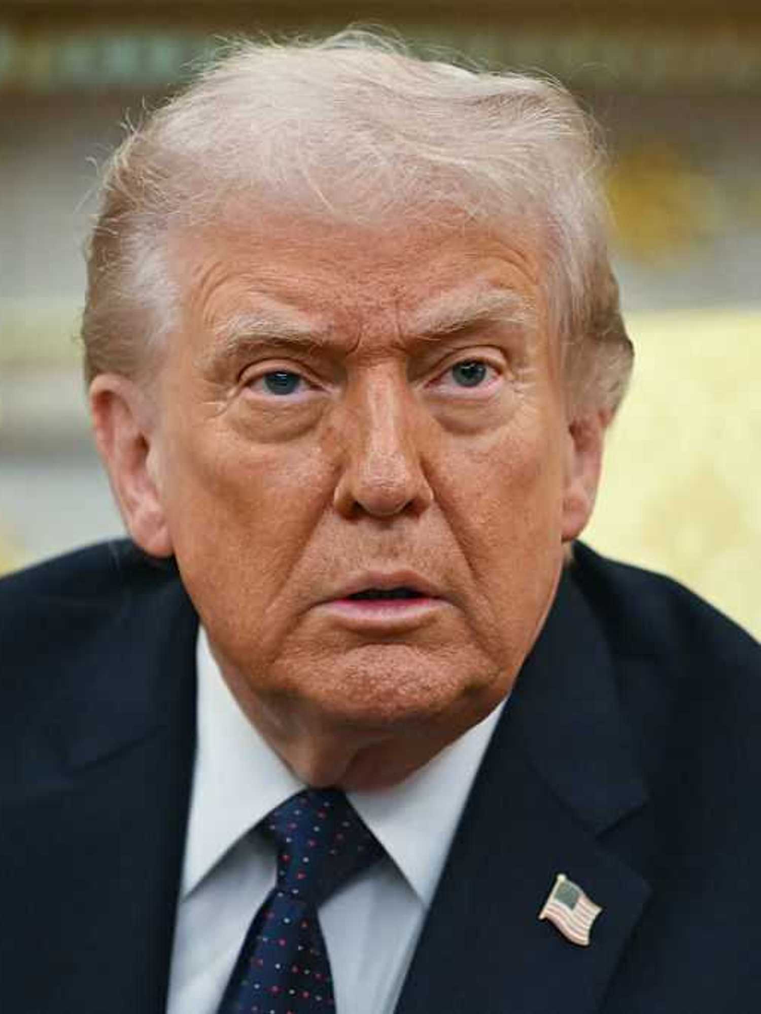 Donald Trump with a neutral expression on his face, sitting on a cream chair in the Oval Office.
